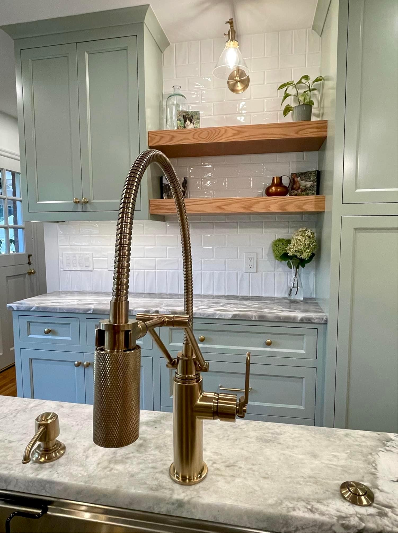 Soft green painted flush-inset kitchen cabinetry with white oak floating shelves, marble countertops and brass fixtures